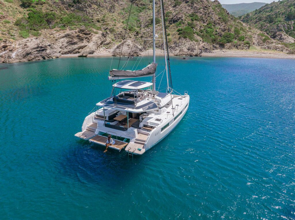Mediterranean catamaran anchorages - Lagoon catamaran peacefully anchored in a protected bay, with bow anchor set and bow facing the pristine beach, surrounded by crystal-clear turquoise waters